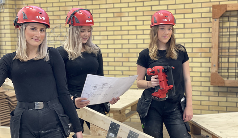 Tre tjejer i bygghjälmar står i en bygghall.